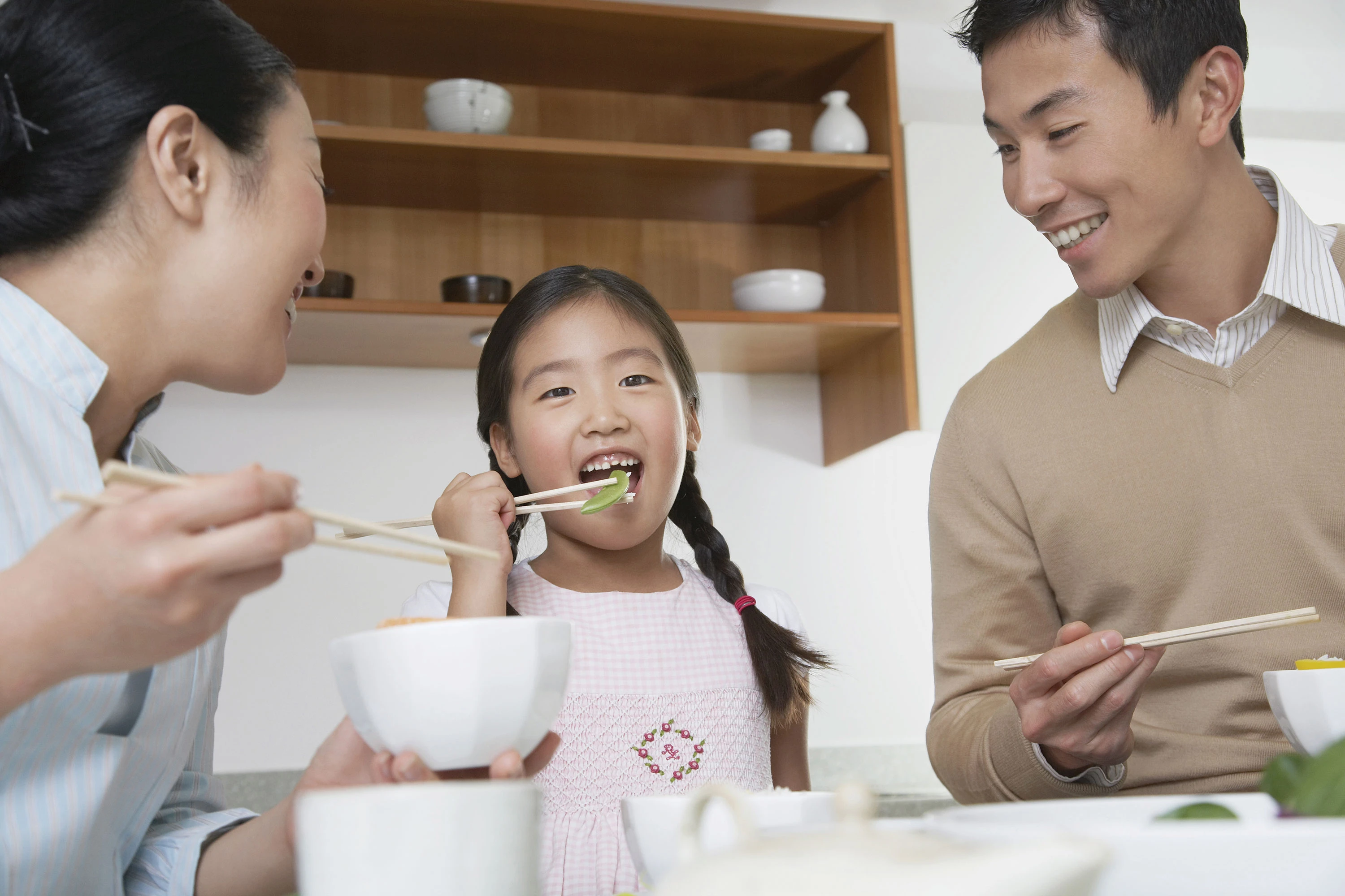 family of three having a meal at home with girl putting vegetable into her mouth