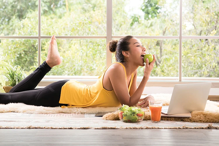 smiling young woman in yellow active wear eating an apple while using laptop